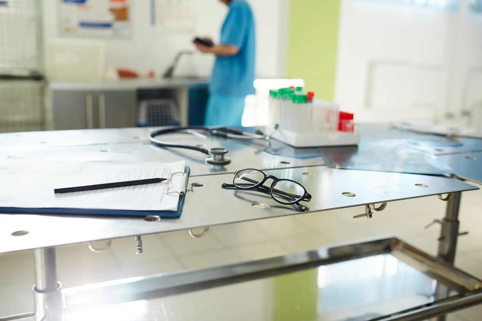 Close-up of stainless steel laboratory table with clipboard, glasses, stethoscope, and test tubes in medical setting